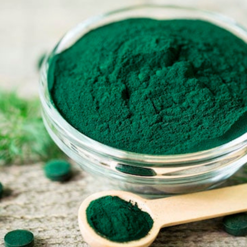 Deep green Spirulina powder in a glass bowl with a small wooden spoon beside it.